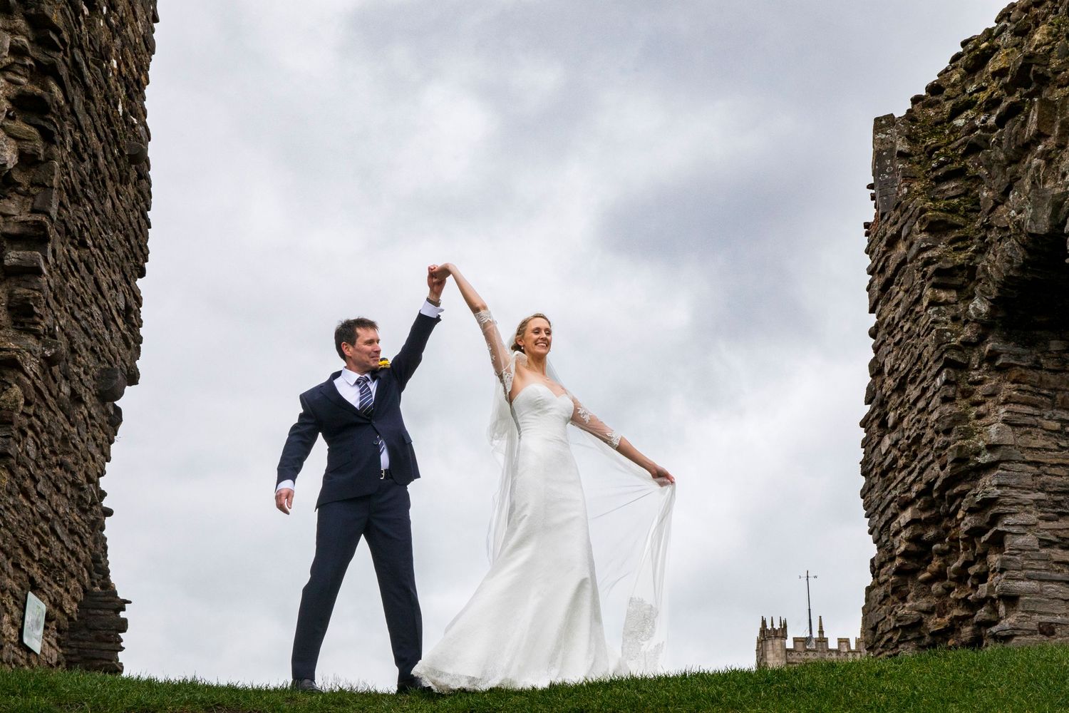 Newlyweds strike triumphant pose between ancient stone ruins against cloudy sky.