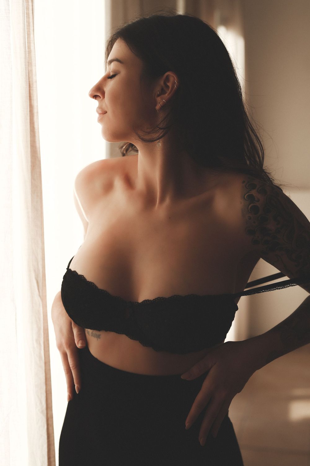 Tattooed woman in off-shoulder black top gazing out window in warm natural light.