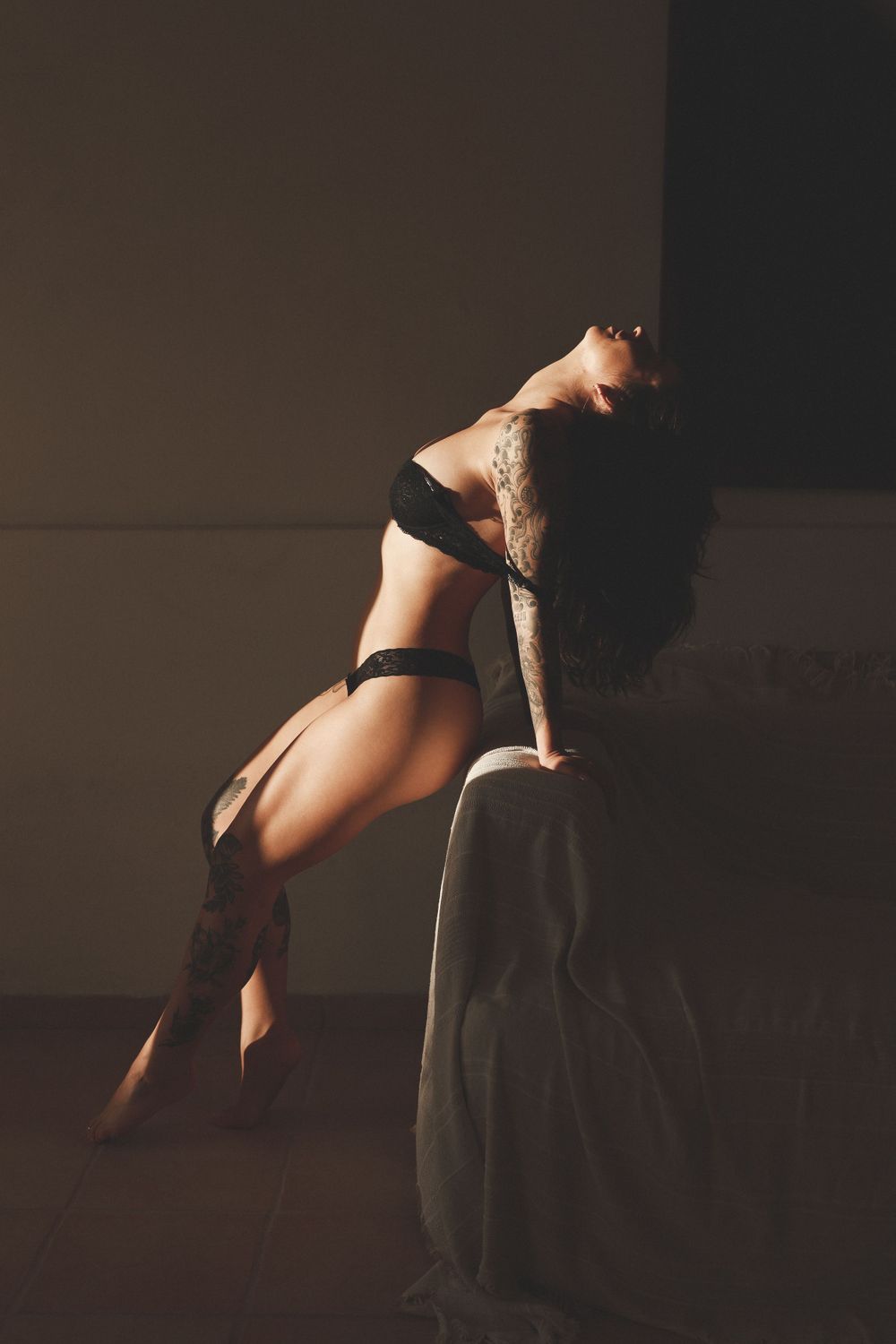 Tattooed woman in black lingerie arching back against wall in dramatic moody lighting.