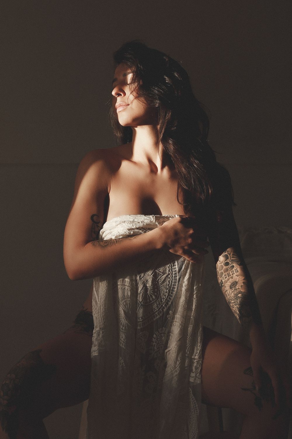 Tattooed woman holding white lace fabric in warm dramatic studio lighting boudoir photo.