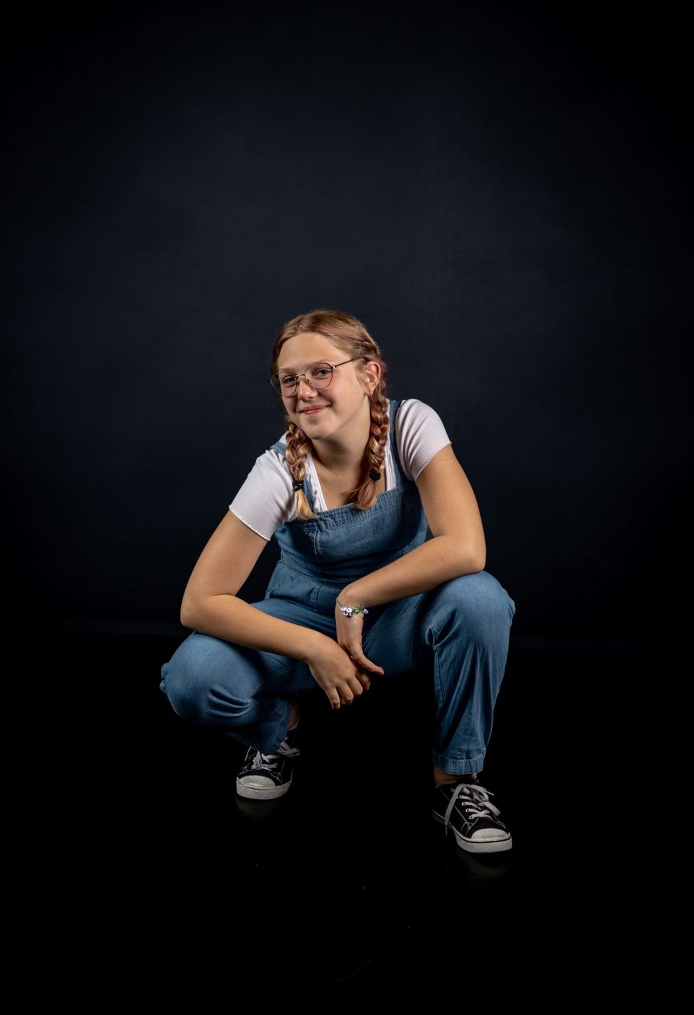Studio portrait in overalls and braids sitting against dark background.