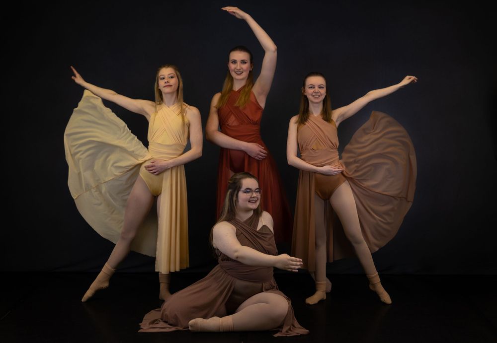 Group of dancers in earth-toned dresses pose dramatically against black backdrop.