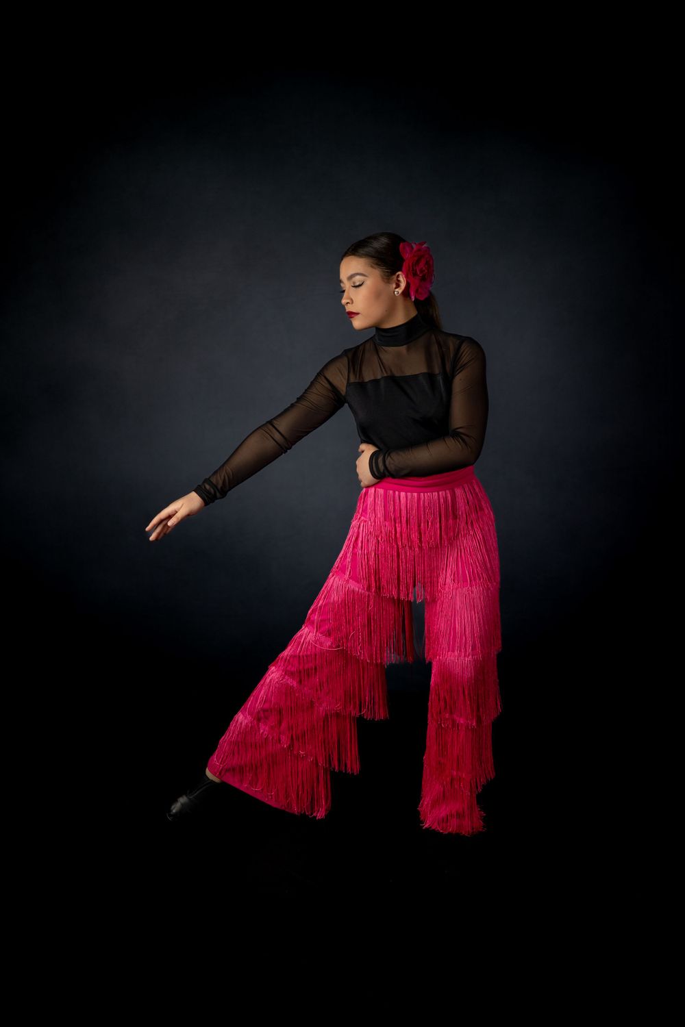 Dancer in sheer black top and flowing red fringe pants poses against dark background.