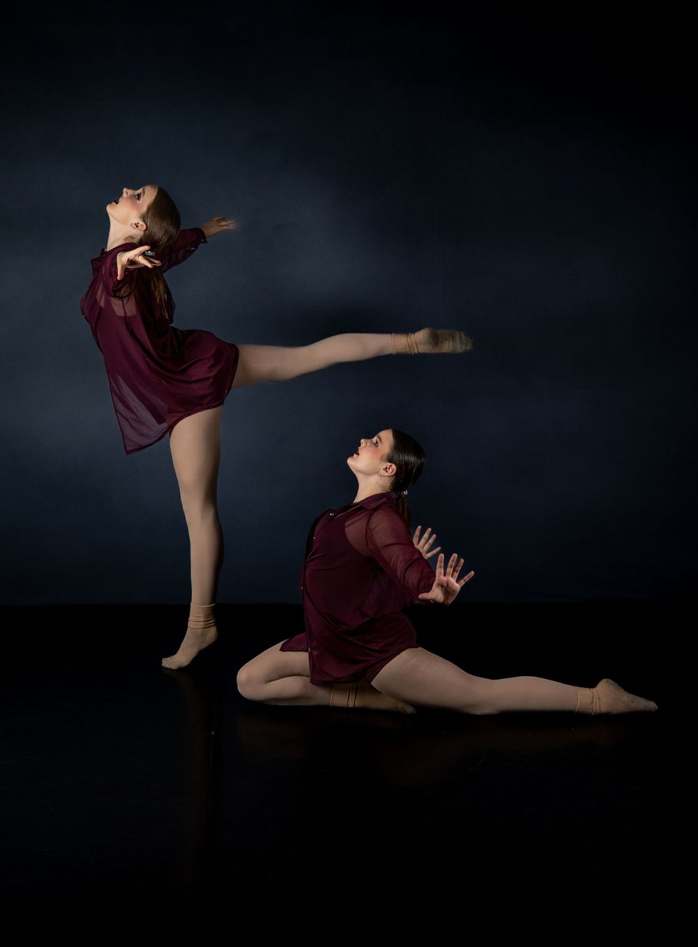 Two dancers in burgundy dresses perform dynamic poses against dark background.