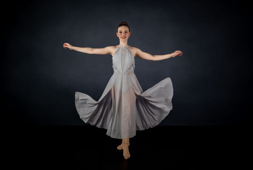Dancer in grey halter dress spins with arms outstretched against black backdrop.