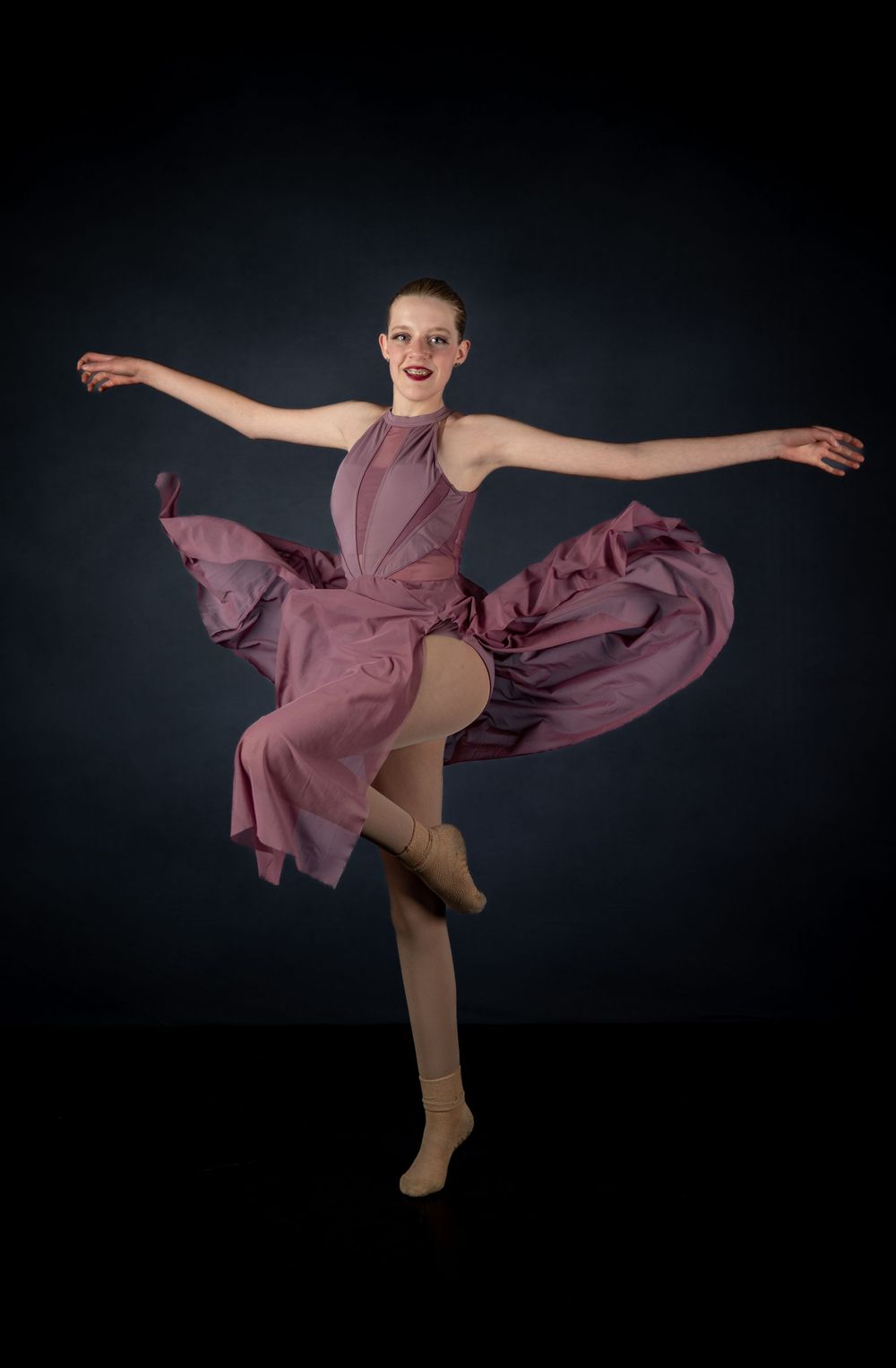 Dancer in mauve dress performs energetic pose with flowing fabric against dark background.