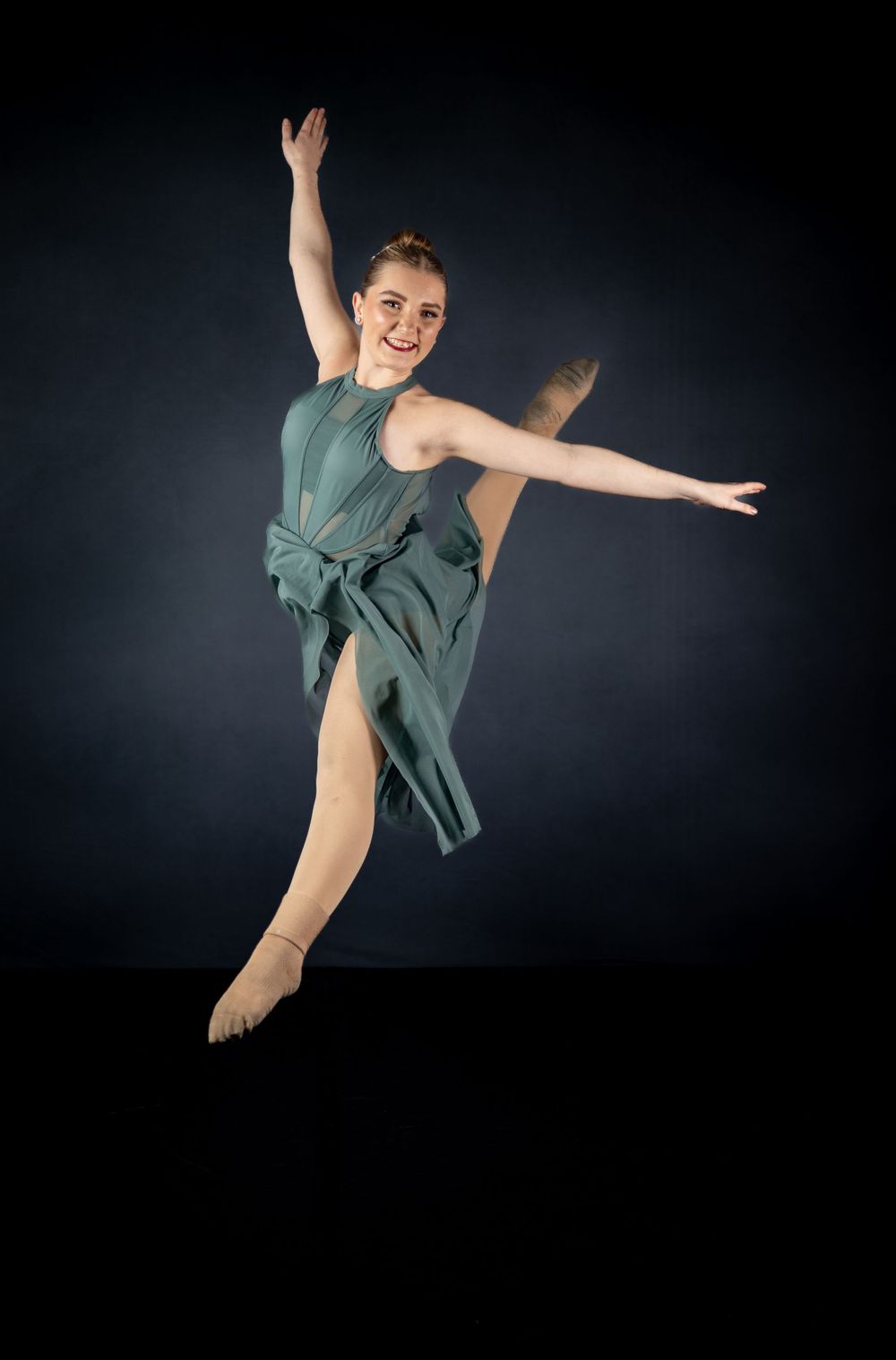 Dancer leaps gracefully in sage green dress against black studio backdrop.