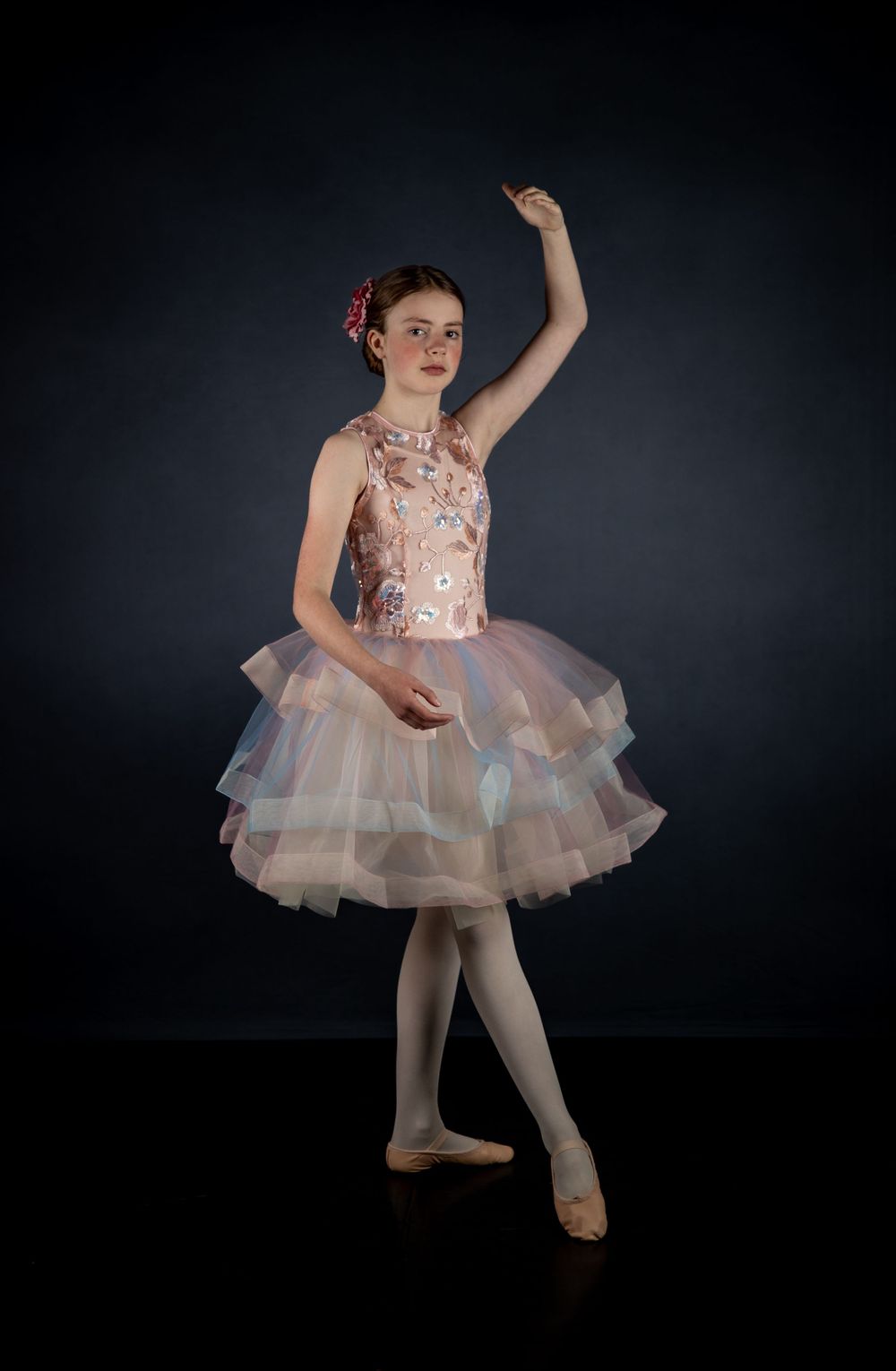 Ballet dancer poses in pink floral tutu dress with layered tulle skirt against dark backdrop.