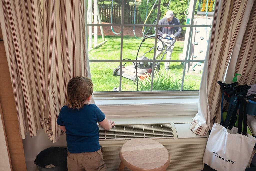 Child looking out window at playground equipment on sunny day with beige curtains framing the view.