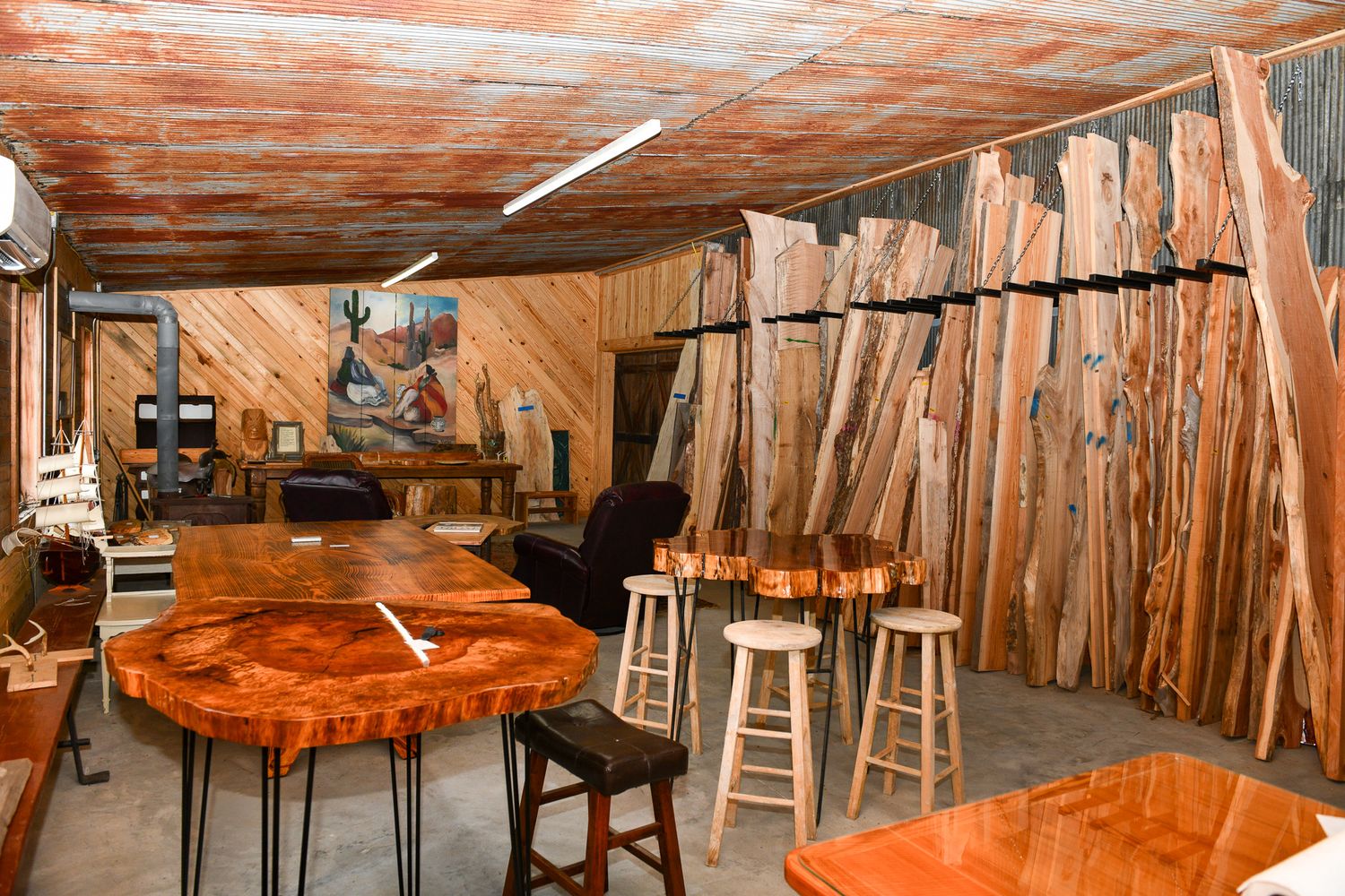 Wood workshop interior showing finished tables and raw wood slabs stored along wall.