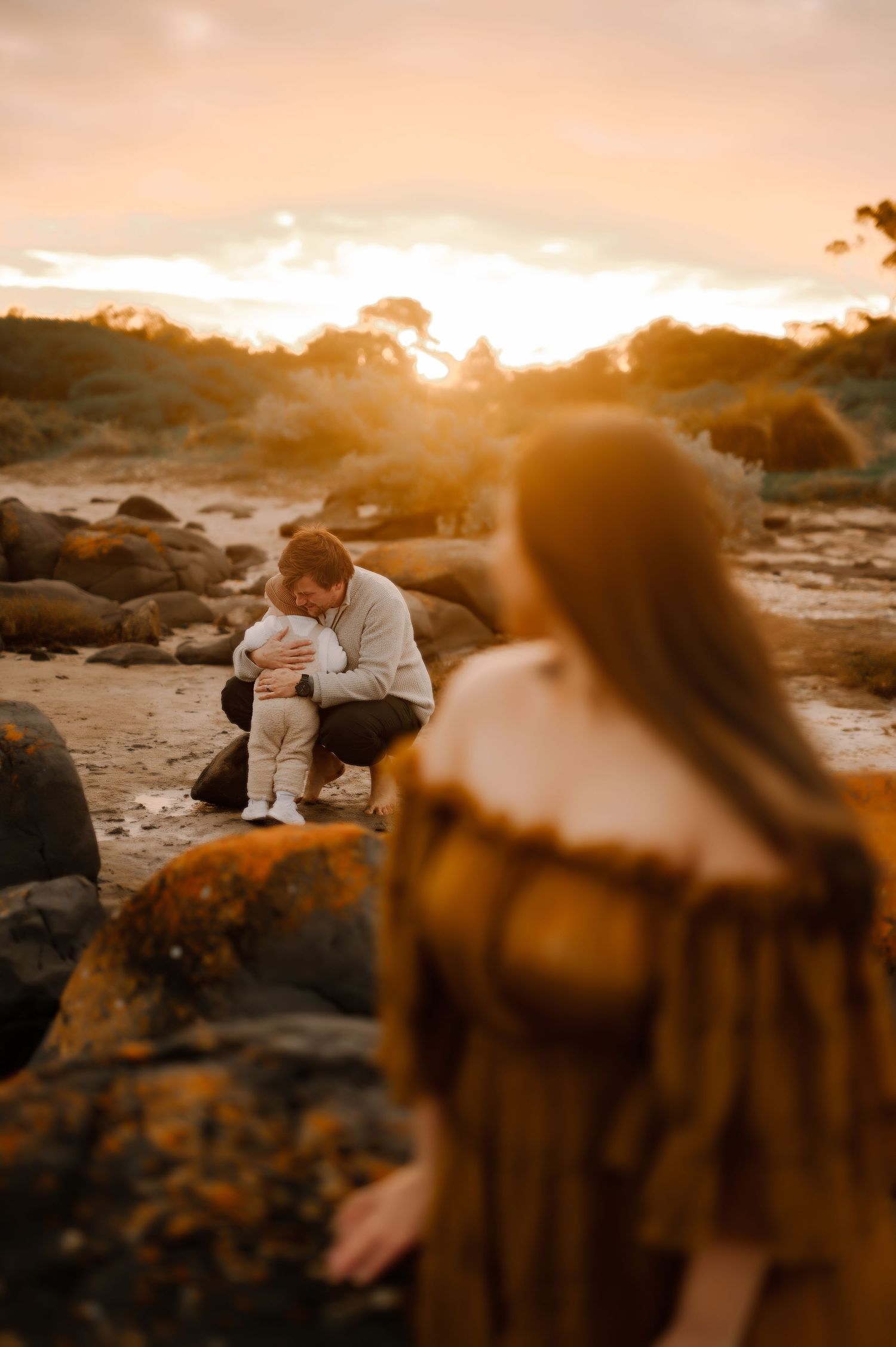 Sunset beach scene with figures among rocky coastal landscape.