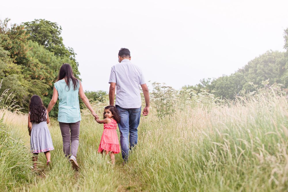 A family walks together through a tall grass meadow on a sunny summer day, photographed from behind.