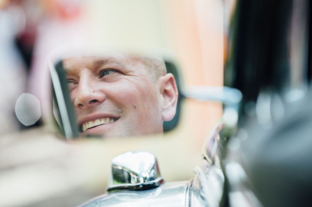 A person smiles while looking at a car's rearview mirror during daytime.