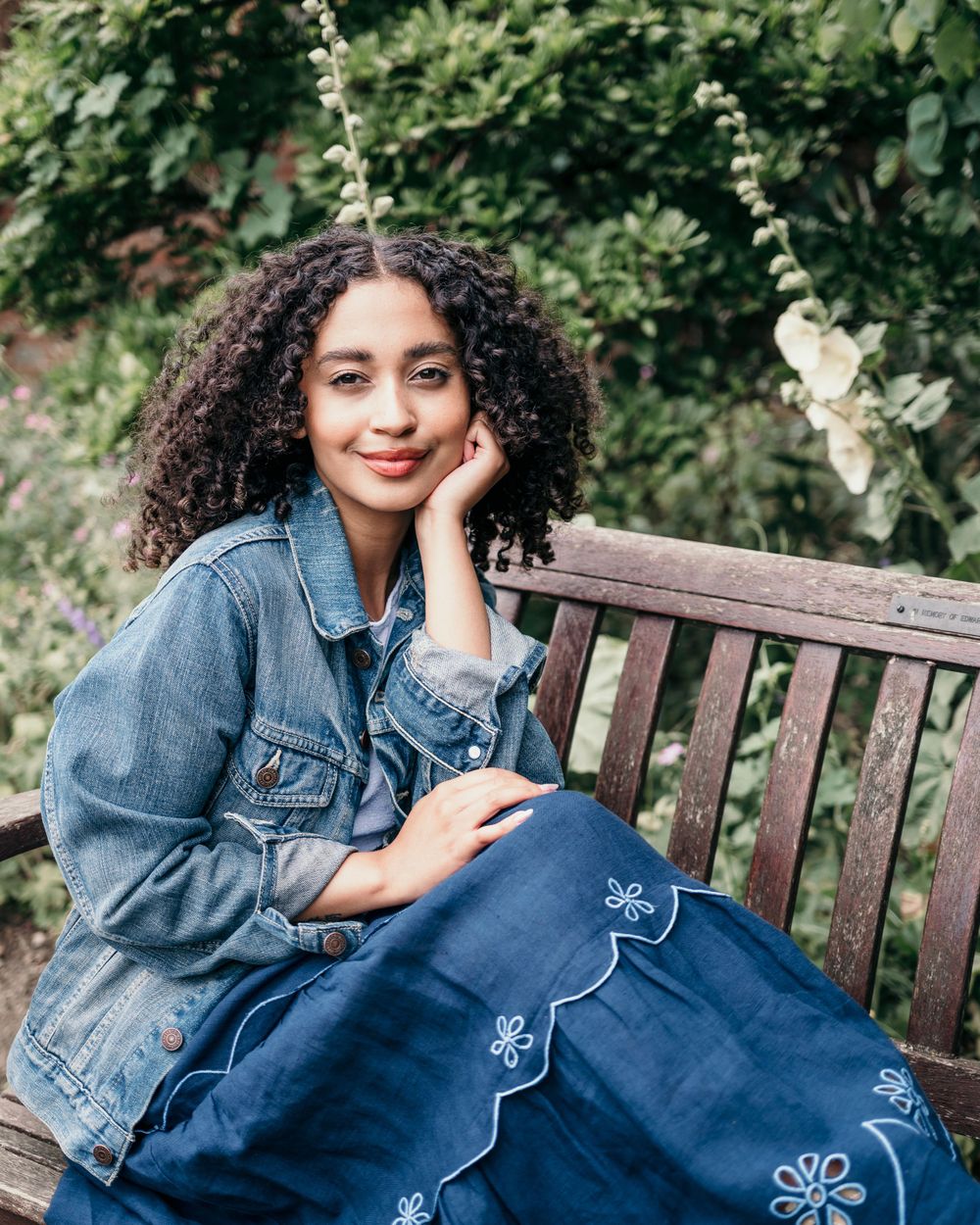A person in a denim jacket relaxes on a wooden bench in a lush garden setting with natural lighting.