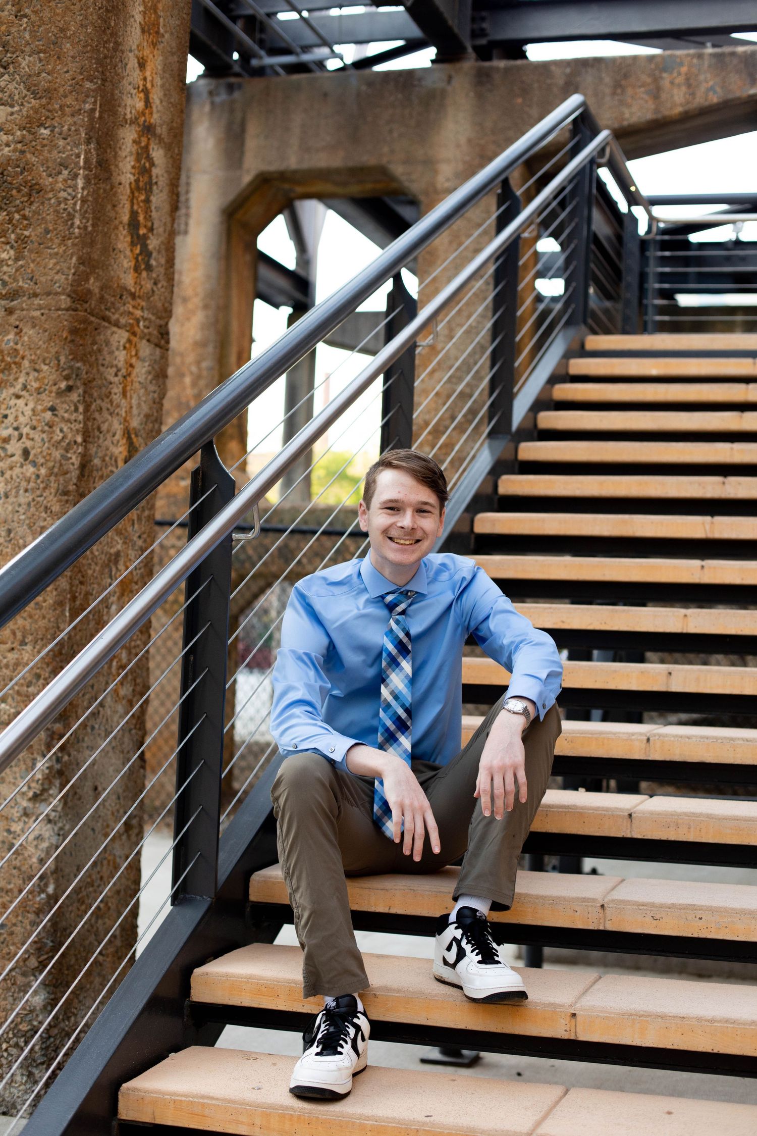 Student in blue shirt and khakis sits casually on modern wooden and metal stairs.