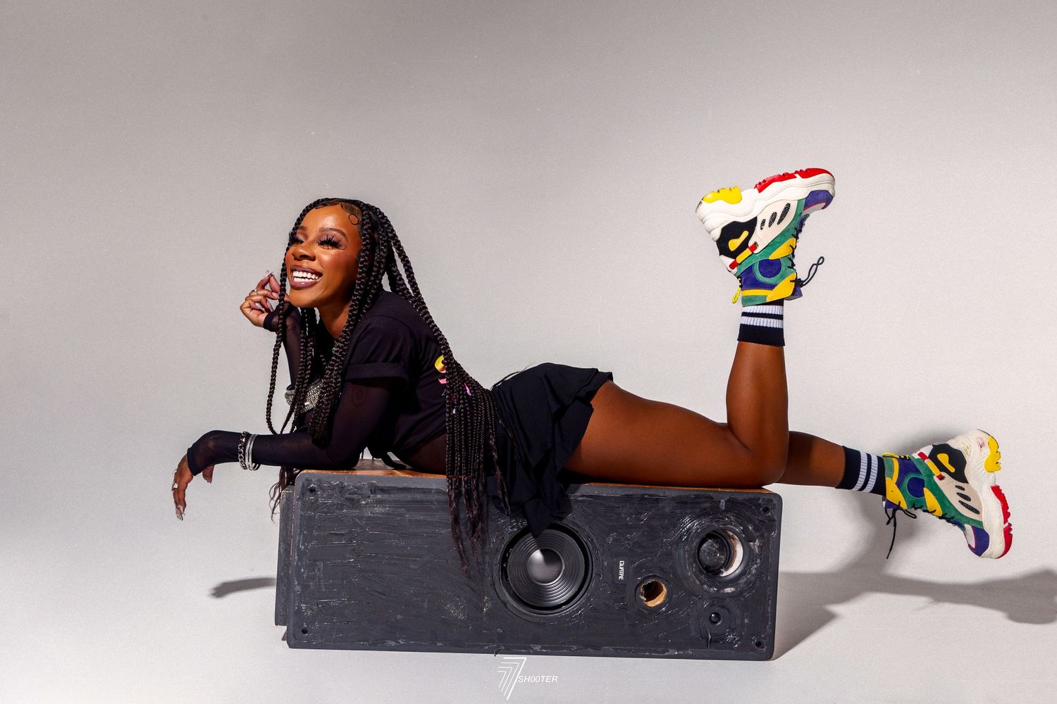 Energetic person poses playfully on a large black speaker wearing colorful sneakers in a studio setting.