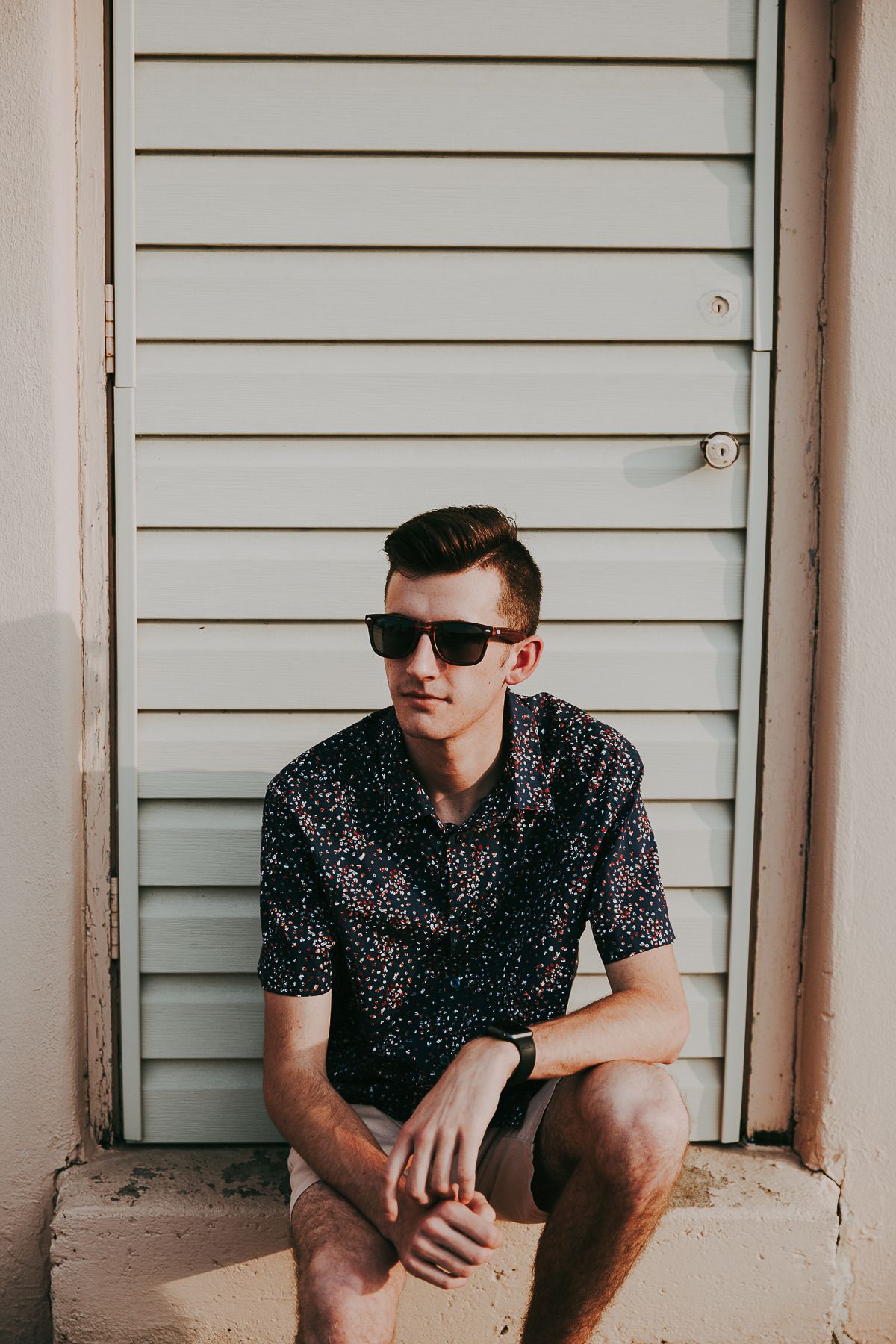 high school senior boy sitting on steps looking to the left in patterned shirt in front of shutters with sunglasses