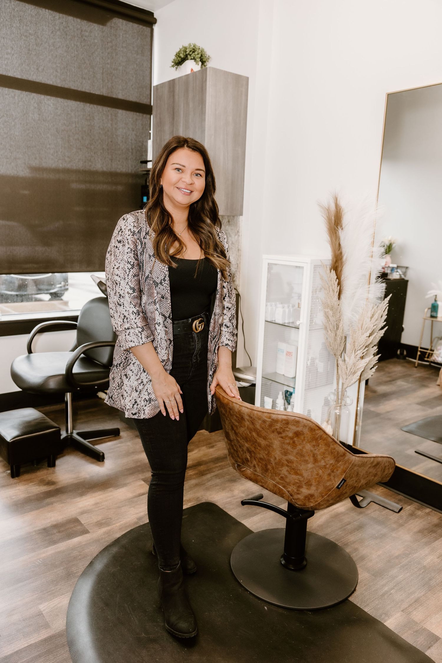 A stylist in snake print blazer stands next to a brown salon chair in a modern salon setting.