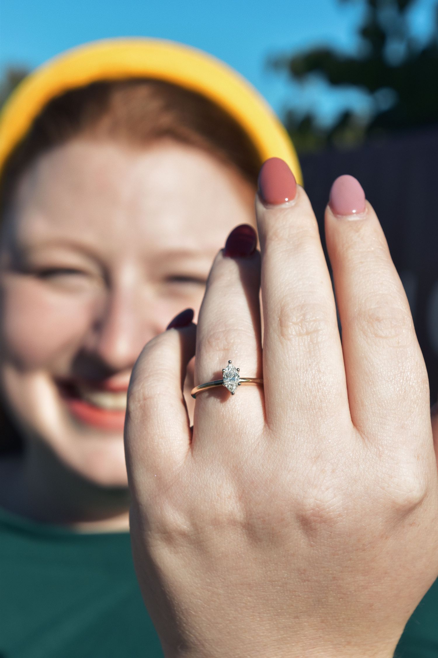A smiling red-haired woman wearing a yellow headband proudly displays a solitaire diamond engagement ring on her finger.