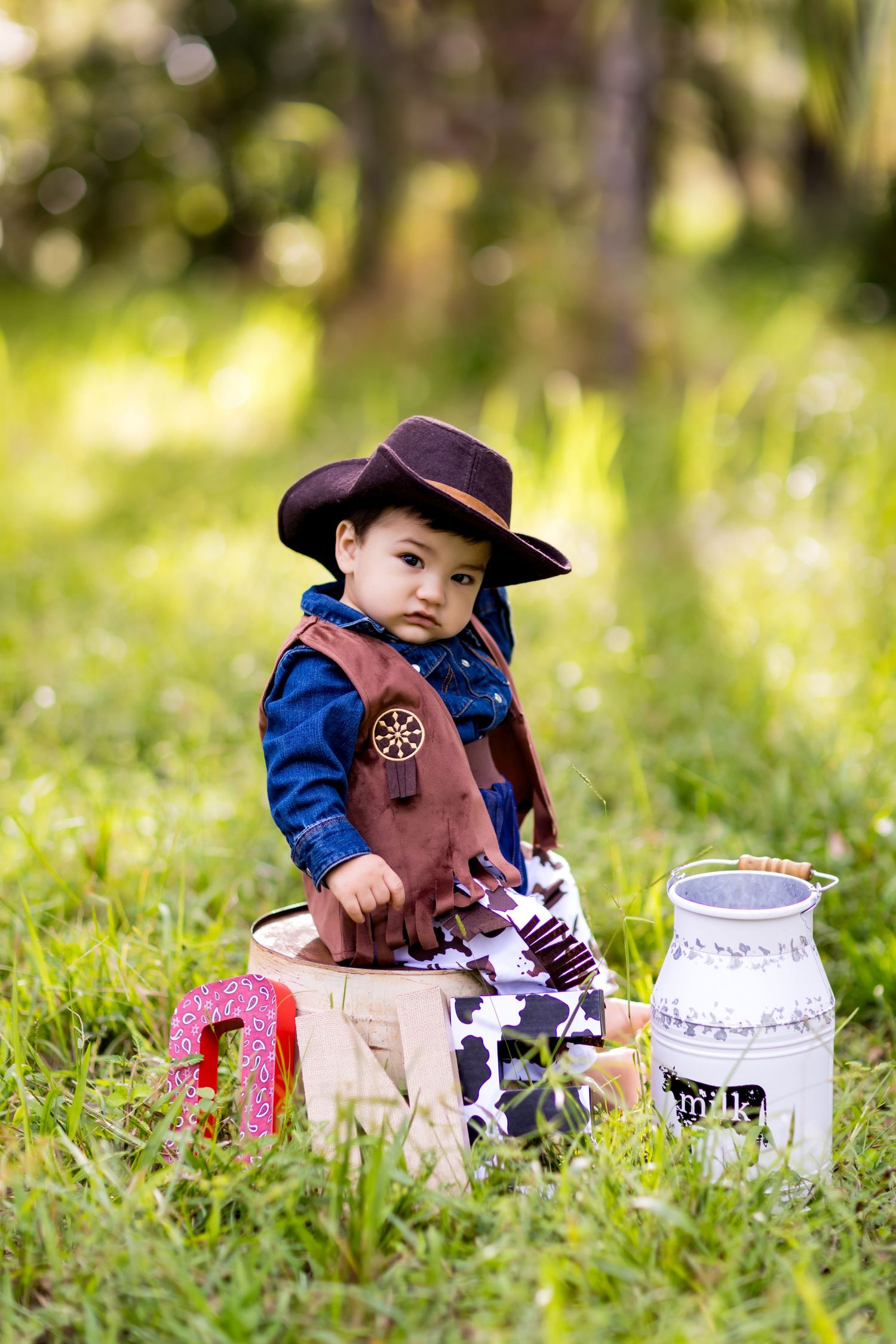 Toddler in cowboy attire sits on wooden crate with milk jug and toys in sunny meadow.