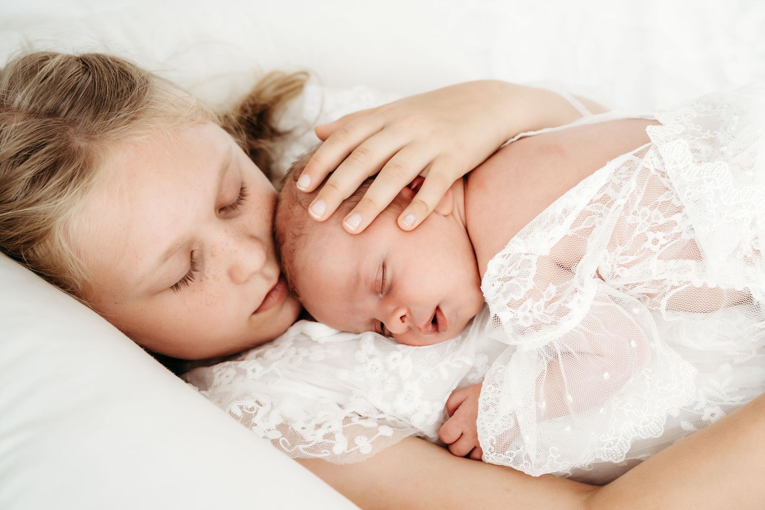 Two sleeping children cuddle together on white bedding with delicate lace details.