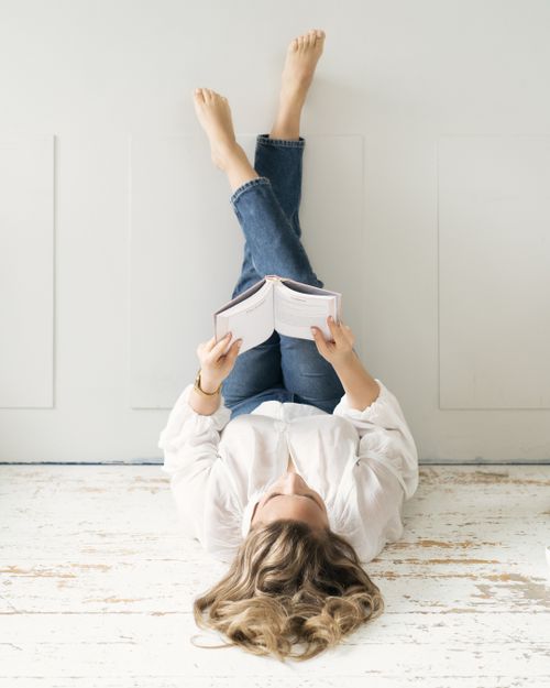 Person in white shirt and jeans lies on floor while reading book with feet up against wall.