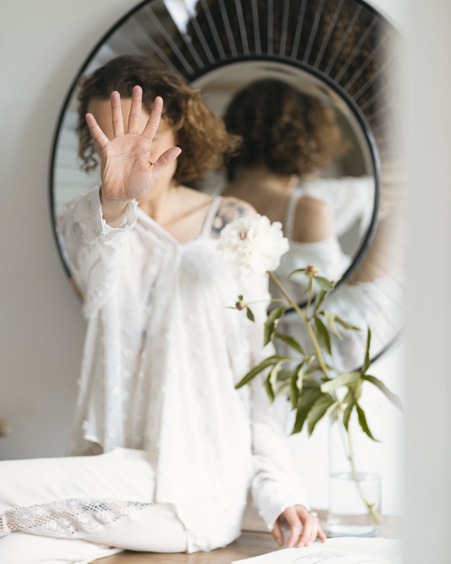 Hand raised in front of sunburst mirror with white clothing and plant decor.