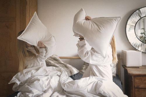 Two people having a playful pillow fight on a bed with white bedding.