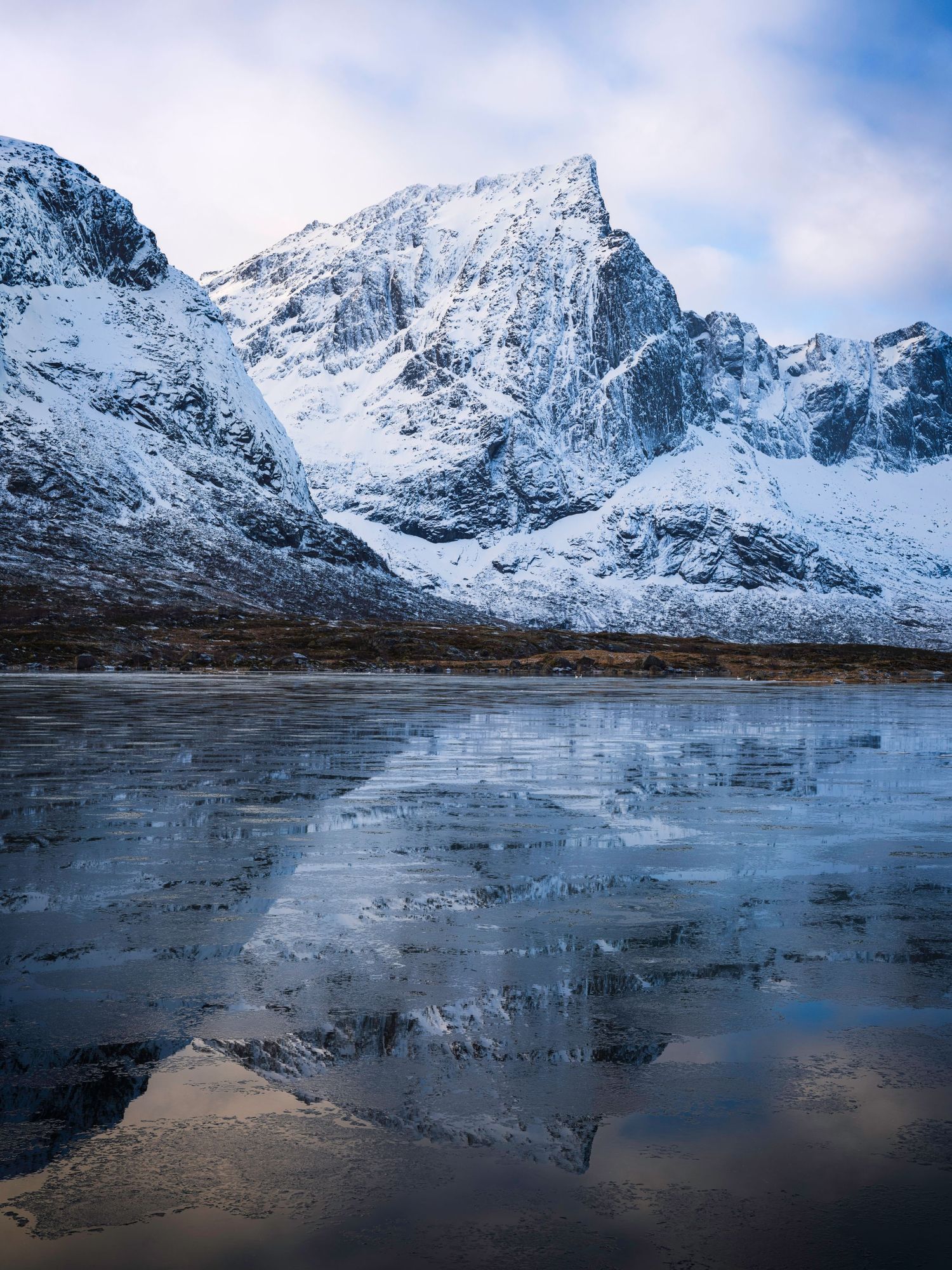 Snow-dusted mountain peaks reflect perfectly in the still waters of a fjord under blue skies.