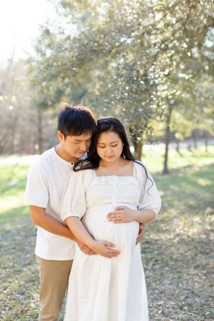 Expectant couple shares tender moment in sunlit park setting.