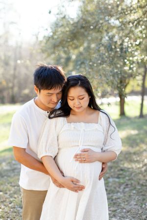Couple embraces while cradling baby bump in natural outdoor setting.