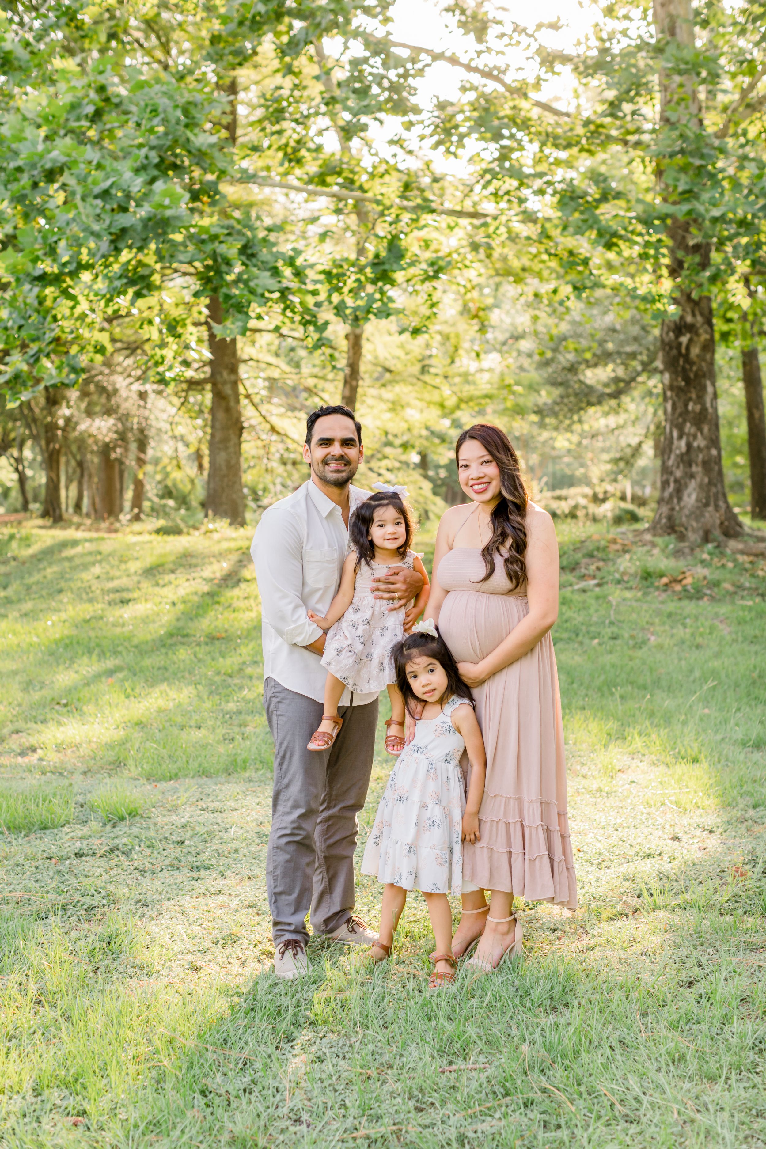 Family of four poses together in sunlit wooded area wearing coordinated neutral outfits.