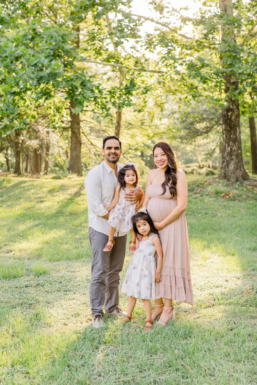 Family of four poses together in sunlit wooded area wearing coordinated neutral outfits.