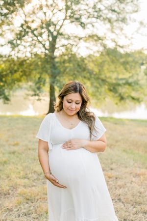 A maternity photo in a sunlit field shows an expectant mother in a flowing white dress tenderly cradling her belly.