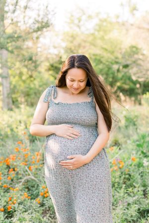 A pregnant woman in a floral dress tenderly holds her baby bump among wildflowers.