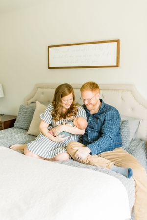 A couple sits on a bed holding their newborn baby in a bright, modern bedroom.