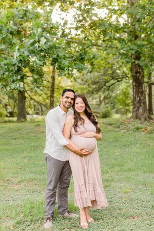 Expecting couple embraces in a sunlit forest setting wearing coordinated neutral outfits.