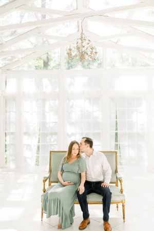 An expectant couple sits together on an antique settee in a bright sunroom.