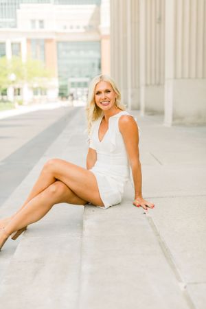 A graduate in white sits on outdoor steps while holding a diploma.
