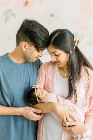 Couple sharing intimate moment while cradling their newborn baby in pink-themed nursery.