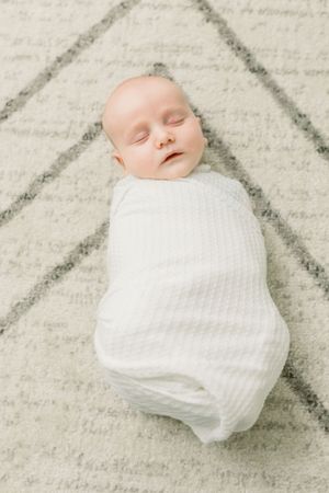 Peaceful sleeping newborn baby wrapped in white swaddle on a patterned rug.