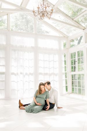 A maternity photoshoot in a bright sunlit conservatory with white wooden beams and a vintage chandelier.