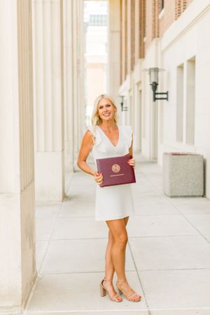 A graduate in a white dress poses with their diploma on a bright outdoor walkway.
