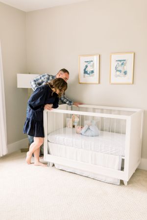 Parents lean over white crib together in modern nursery with wall art.