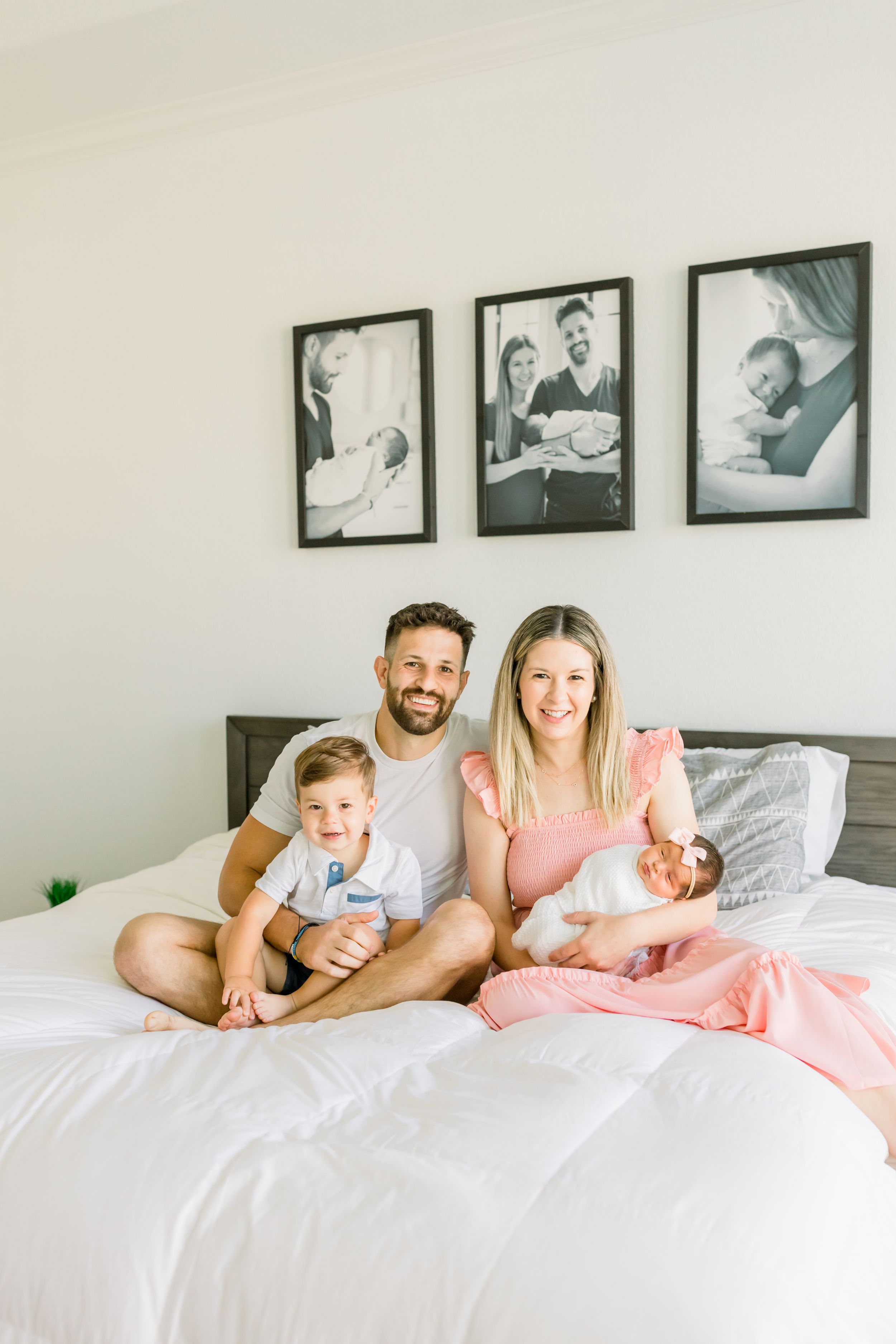 Family poses on bed with framed photos displayed on wall behind them.
