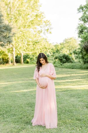 A maternity photo of a woman in a flowing pink dress standing on grass in golden sunlight.