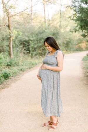 An expectant mother in a floral dress tenderly touching her baby bump on a sunlit path.