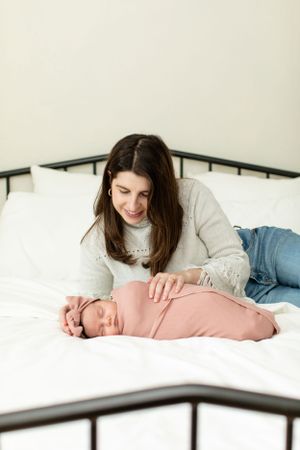 Mother lying on bed beside sleeping newborn baby in pink wrap.