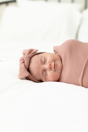 Peaceful sleeping newborn wearing pink knit wrap and bow on white bedding.