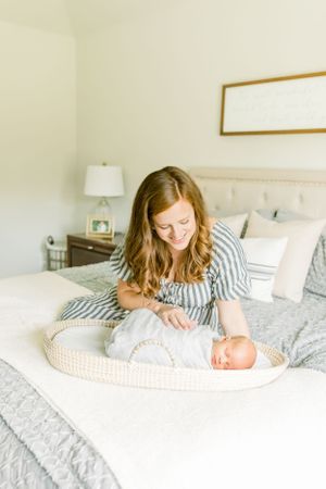 A woman in striped dress tends to her newborn baby lying in a woven basket on the bed.